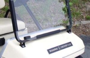 Clear one-piece windshield installed on a golf cart, showing the transparent pane, hinges, and lower mounting brackets.