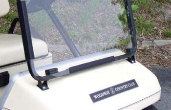 Clear one-piece windshield installed on a golf cart, showing the transparent pane, hinges, and lower mounting brackets.